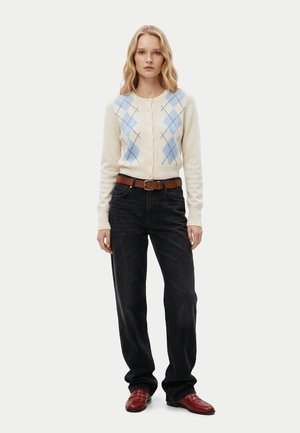 Une femme porte un cardigan crème avec un motif losanges bleu, un jean noir avec une ceinture marron et des chaussures en cuir rouges, debout devant un fond uni.