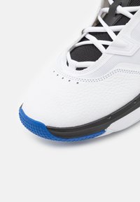 Biały sneaker z teksturowaną skórzaną cholewką, czarnym językiem z siateczki oraz niebieskimi gumowymi akcentami. Posiada czarną śródpodeszwę i białe sznurówki.