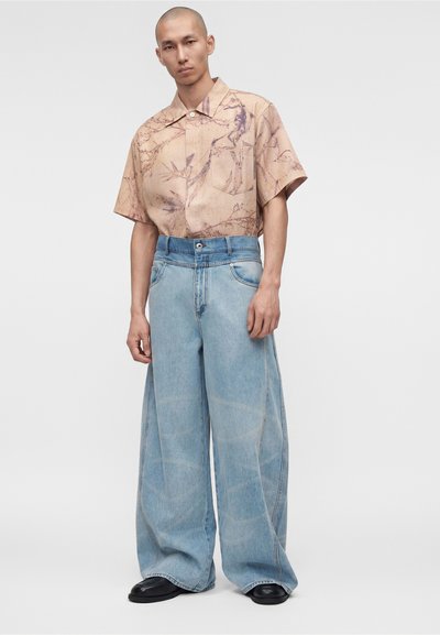 Άντρας που φοράει μπεζ κοντομάνικο πουκάμισο με αφηρημένο σχέδιο, ανοιχτόμπλε τζιν φαρδιάς γραμμής και μαύρα παπούτσια, στέκεται μπροστά σε ένα μονόχρωμο ανοιχτό φόντο.