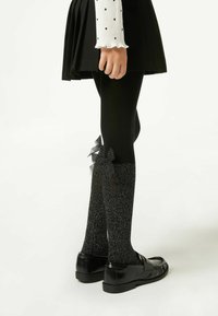 Czarne legginsy z błyszczącym wykończeniem, z teksturowanym, błyszczącym górnym elementem i czarnymi kokardkami przy mankietach, w połączeniu z błyszczącymi czarnymi butami.