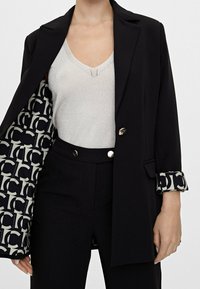 Czarna marynarka z złotymi guzikami, z wzorzystym wnętrzem z literami, noszona na srebrnej bluzce i czarnych spodniach. Podwinięte rękawy odsłaniają wzór.