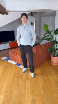 Grijze trui met ritssluiting, zwarte broek en zilveren sneakers. Kamer heeft een houten vloer, een oranje bank en een grote groene plant.