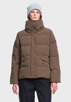 Jeune personne portant une doudoune marron avec un col montant et des poches zippées, un bonnet tricoté gris clair et un pantalon foncé.