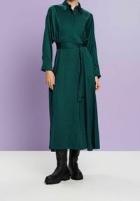Robe à manches longues, couleur vert émeraude, avec col et ceinture à la taille, fabriquée en tissu lisse. Associée à des bottes noires montantes jusqu'au genou, à semelles épaisses.