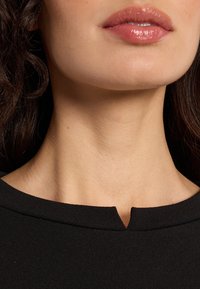 Close-up of a person’s lower face and neck with glossy pink lips, wearing a black top with a small notch at the neckline.