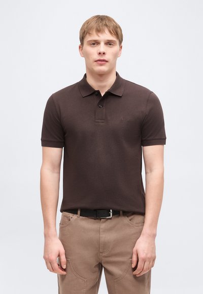 Jeune homme portant un polo à manches courtes marron foncé et un pantalon beige, debout devant un fond clair uni.