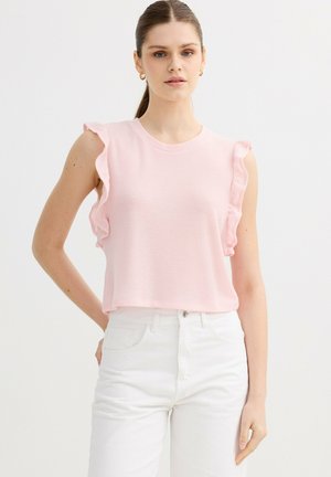 Mujer con un top sin mangas de color rosa claro con bordes con volantes combinado con pantalones blancos de cintura alta, posando frente a un fondo liso.