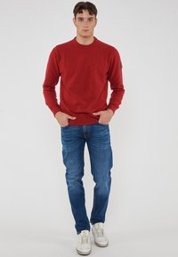 Giovane uomo in piedi con le mani in tasca, indossando un maglione rosso, jeans blu e sneakers bianche, su uno sfondo semplice e chiaro.