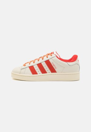 Witte lage sneaker met oranje strepen, veters en binnenvoering, met het label "Campus" aan de zijkant, platte zool, tegen een neutrale achtergrond.