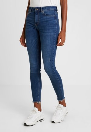 Kvinde iført blå skinny jeans med mellemhøj talje og frynser ved kanten samt hvide sneakers, stående mod en ensfarvet lys baggrund.
