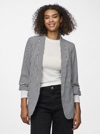 Fekete-fehér houndstooth mintás blézer felgörgő ujjakkal, világos felsőre húzva, fekete nadrággal párosítva. Simának tűnő szövettextúra.