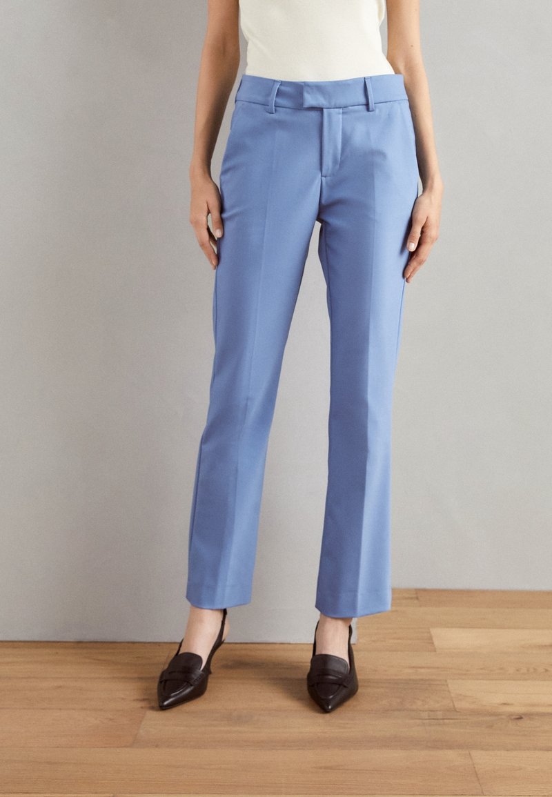 Světle modré kalhoty na míru se střihovým střihem, předními záhyby a dvěma postranními kapsami. Pár je doplněn tmavě hnědými botami s špičatou špičkou.
