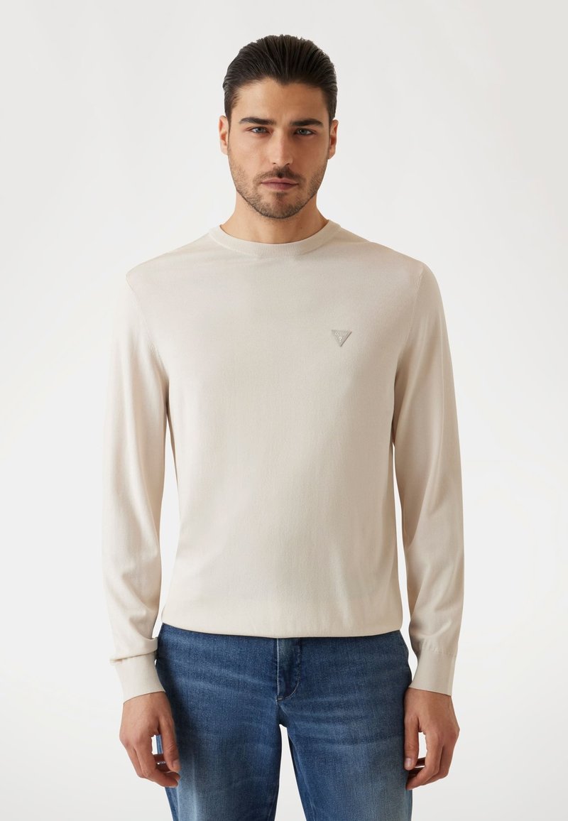 Camicia beige a maniche lunghe in morbido tessuto con scollatura a barca, dotata di un piccolo logo sul petto a sinistra. Abbinata a jeans blu.