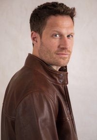 Man with short brown hair wearing a brown leather jacket looks sideways against a plain beige background.