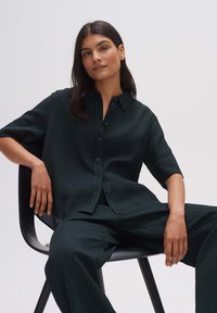 Dark green, textured shirt with a button-up front and short sleeves, paired with matching textured pants. Model seated on a black chair.