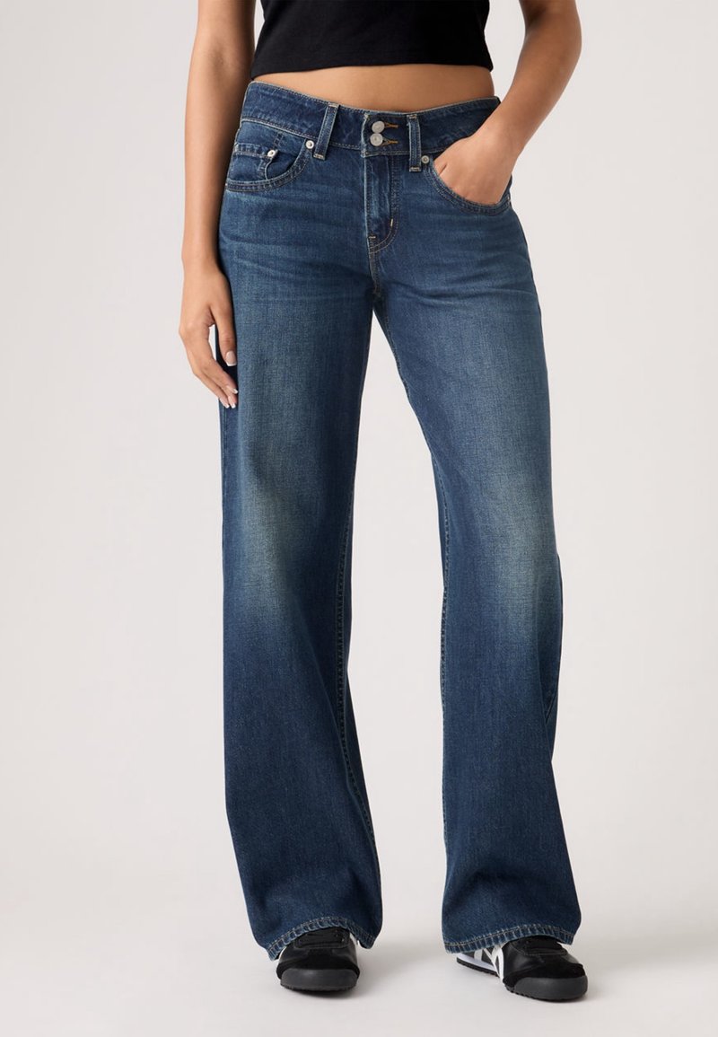 Femme portant un jean bleu en denim taille haute à jambes larges avec poches avant, associé à des baskets noires et un top court noir.