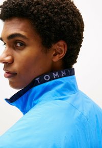Lichtblauwe windjack met een hoge kraag en een donker marineblauwe bies met de tekst "TOMMY" in vetgedrukte letters. Getextureerde stof met zichtbare stiksels.