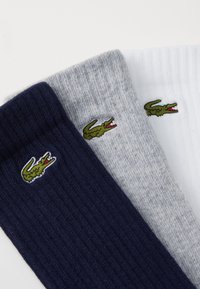 Drei Paare gestreifter Socken: marineblau, grau und weiß, jeweils mit einem kleinen grünen und roten Krokodil-Logo nahe der Oberseite.