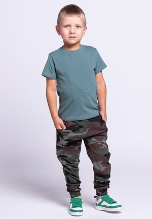 Młody chłopiec stoi z rękami w kieszeniach, ubrany w zielony t-shirt, spodnie w kamuflaż oraz zielono-białe sneakersy na jednolitym tle.