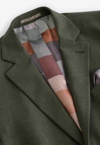 Blazer vert en tissu texturé, avec une doublure en satin multicolore à rayures. Un mouchoir de poche est visible dans la poche avant.