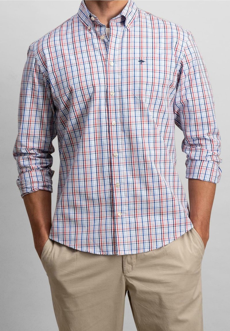 Homme portant une chemise à carreaux rouge, blanc et bleu avec les manches retroussées et un pantalon beige, les mains dans les poches.