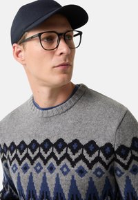 Junger Mann mit schwarz gerahmten Brille, marineblauem Hut und grauem Pullover mit blauen und schwarzen geometrischen Mustern, der leicht nach rechts schaut.