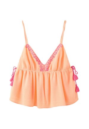 Jeune femme debout portant un haut sans manches couleur pêche avec broderie rose, une jupe en jean blanche et des sandales blanches à enfiler sur fond uni.