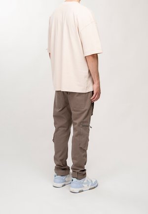 Camiseta de algodón beige de corte oversized, pantalones cargo marrones con cremalleras laterales y zapatillas de deporte azul claro. Diseño sencillo, ajuste relajado y textura suave.