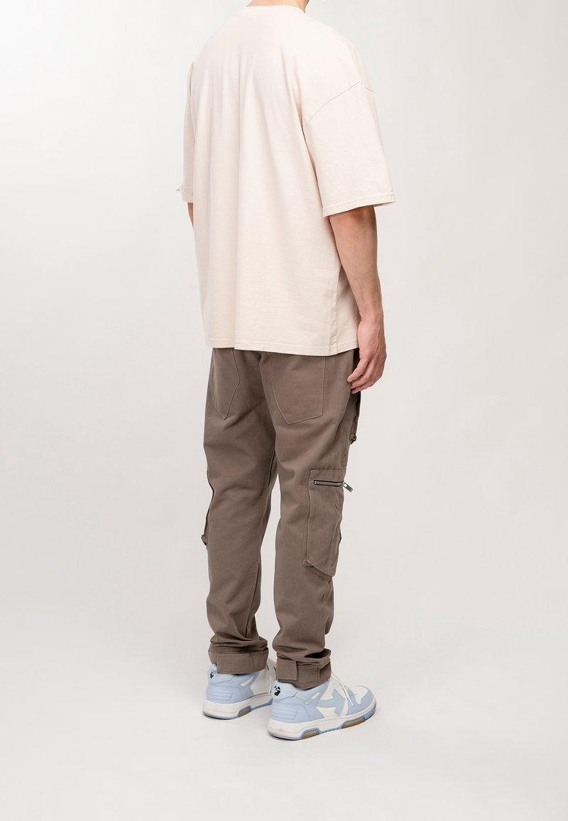 Beige Baumwoll-Übergrößen-T-Shirt, braune Cargo-Hosen mit seitlichen Reißverschlüssen und hellblaue Sneakers. Einfaches Design, lockere Passform und weiche Textur.