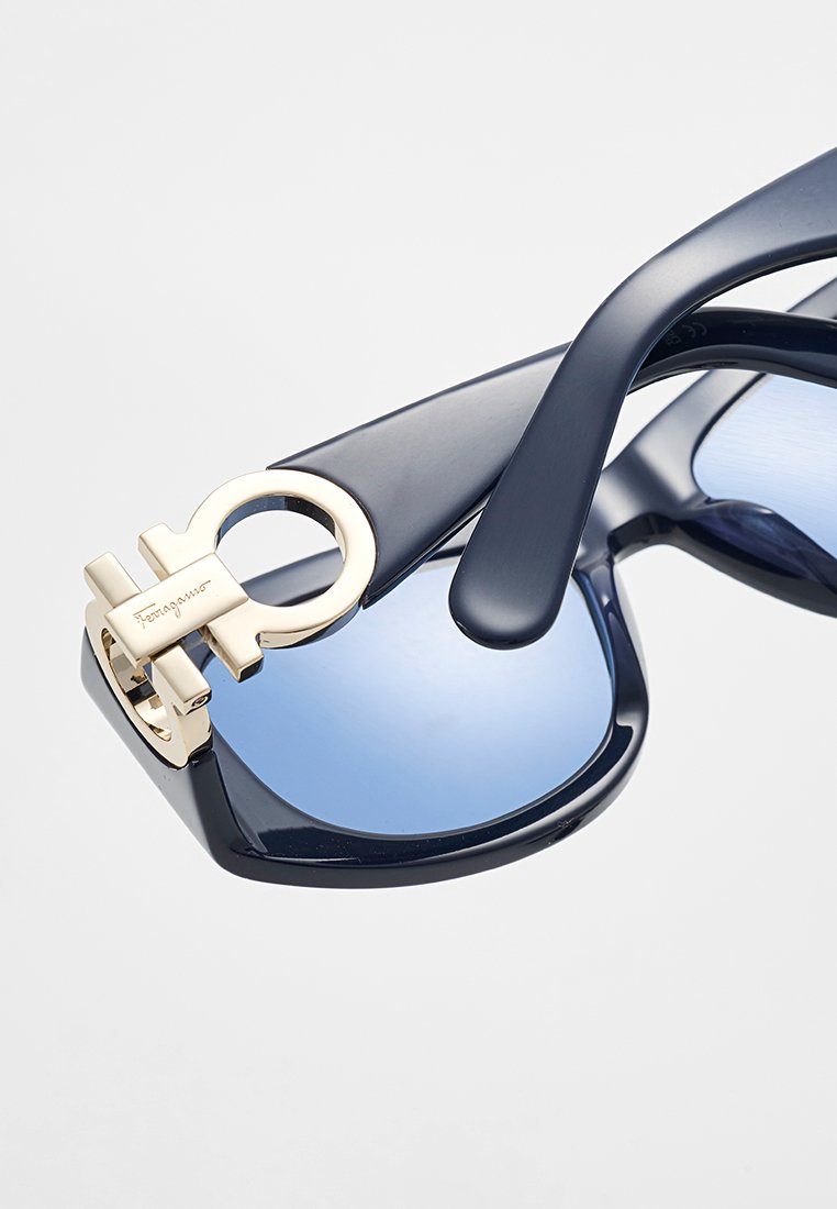 Marinefarbene Acetat-Sonnenbrille mit hellblauen Gläsern. Metallbeschläge in goldener Formakzent an den Bügeln. Glänzende Textur mit glatten Kanten.