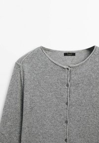 Cardigan gris tricoté avec encolure ronde et cinq boutons noirs sur le devant, montrant la partie supérieure et les détails des épaules.