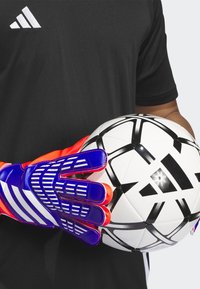 Guanti da portiere Adidas blu con dettagli rossi, dotati di dita con texture antiscivolo, che tengono in mano un pallone da calcio bianco e nero con motivi geometrici.