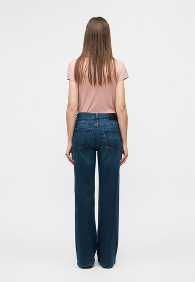 Ροζ ανοιχτό ροζ τοπ με κοντά μανίκια, μπλε τζιν παντελόνι με φαρδιά ποδιά, πίσω τσέπες και λεπτομέρεια με μαύρη ετικέτα, σε προβολή από πίσω πάνω σε απλό φόντο.