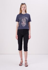 T-shirt grigia con la scritta "SAVAGE" e una grafica di leopardo, abbinata a pantaloni neri cropped e sandali neri con cinturini. Occhiali da sole semplici sulla testa.