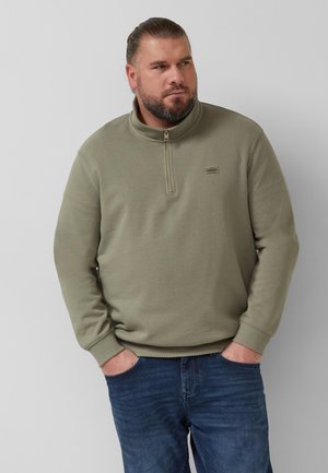 Mężczyzna z brodą w oliwkowym swetrze z zamkiem do połowy i niebieskich dżinsach, stojący z rękami w kieszeniach, patrzący na bok.