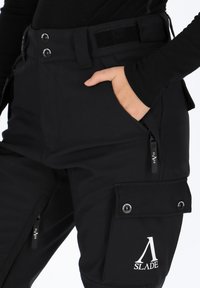 Pantaloni softshell neri con una vestibilità rilassata, dotati di diverse tasche con cerniera e bottone, e un logo del marchio sulla coscia. Vita regolabile.