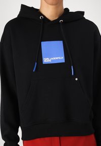 Svart hettegenser med frontlomme, med et blått firkantet logomerke som leser "KARL LAGERFELD JEANS" og blå detaljer på snøringene.