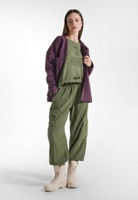 Olivgrüne Cargo-Hosen mit elastischem Bund, kombiniert mit einem figurbetonten olivfarbenen Shirt mit schwarzem Text und einem lila Überhemd. Beigefarbene Kampfstiefel.