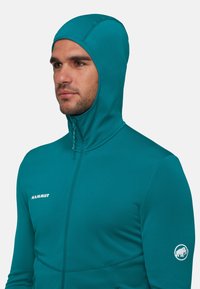 Aansluitende teal fleecejack met een hoge kraag en aangehechte capuchon. Voorzien van een frontzipper en logo op de bovenarm. Zachte textuur.