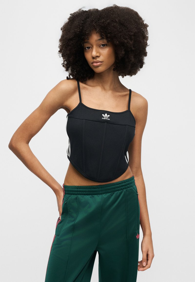 Jeune femme aux cheveux bouclés portant un débardeur noir Adidas et un pantalon de survêtement vert, debout avec une main sur la hanche.