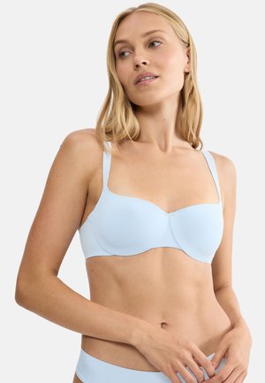 Vrouw met blond haar, gekleed in een lichtblauwe beha en bijpassend ondergoed, kijkt iets naar de zijkant tegen een effen achtergrond.