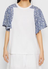 Camisa blanca de mangas cortas con acentos en un patrón floral azul en las mangas. Escote redondo y textura suave. Corte casual con un diseño relajado.