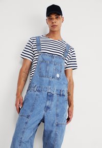 Salopette en denim bleu clair, dotée de bretelles réglables, de poches avant et d'une coupe ample, portée sur un t-shirt à rayures noir et blanc.