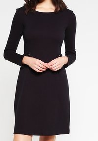 Svart långärmad klänning med rund halsringning, texturerat tyg och dekorativa metalldetaljer vid midjan. Enkel, figurnära A-linjeform.