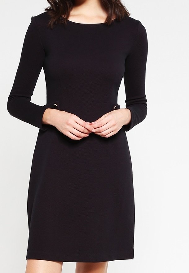 Svart långärmad klänning med rund halsringning, texturerat tyg och dekorativa metalldetaljer vid midjan. Enkel, figurnära A-linjeform.