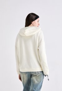 Kremfarget hettegenser med avslappet passform. Mykt stoff, ribbestrikket nederkant og mansjetter. Kombinert med lyseblå jeans med vertikale striper.