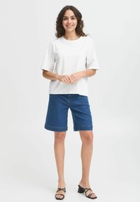 T-shirt bianco a maniche corte, pantaloni bermuda in denim blu e sandali con tacco a blocco neri. Il modello è in piedi con le mani incrociate, sorridendo su uno sfondo chiaro.