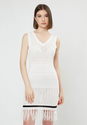 Robe tricotée blanche avec un décolleté en V, présentant un motif ondulé et des rayures horizontales noires. L'ourlet est orné de franges pour plus de texture.
