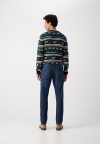 Gebreide trui met een multikleurig patroon van bomen en dieren, gecombineerd met donkerblauwe jeans en beige schoenen. Effen achtergrond.