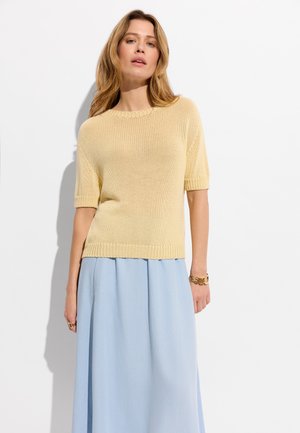 Kobieta o długich blond włosach ubrana w beżowy krótki sweter z dzianiny i jasno niebieską spódnicę midi stojąca na białym tle.